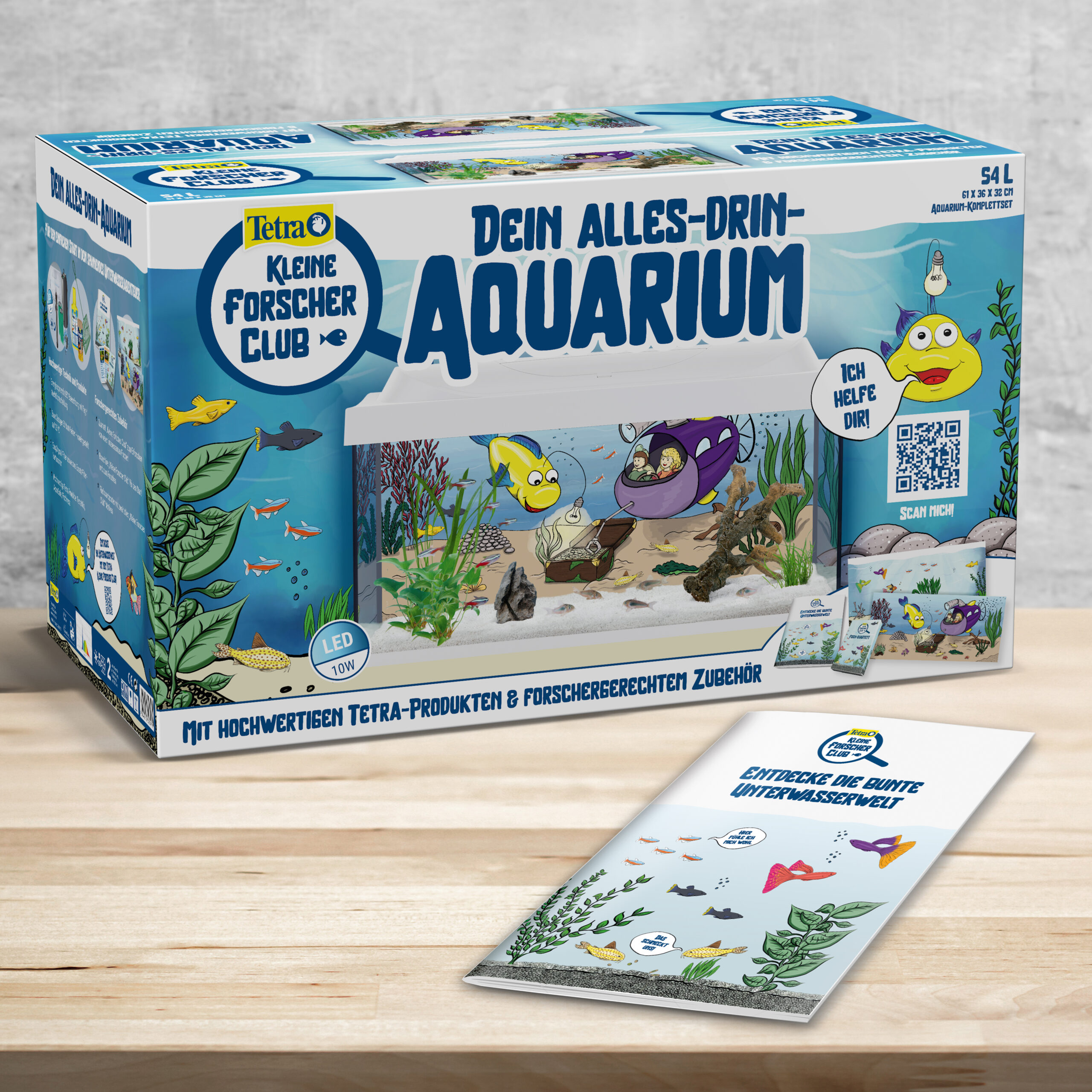 Ein Verpackung des Tetra Kleine Forscher Club "Alles-Drin-Aquarium" steht auf einem hellen Holztisch. Davor liegt eine Broschüre mit kindgerechten Illustrationen zum Thema Aquaristik. Im Hintergrund ist eine Betonwand zu sehen.